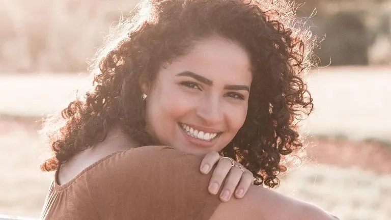 A close-up portrait of a smiling woman with curly hair in sunlight